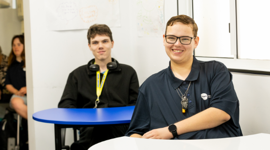 Two STEPS Pathways College students seated at classroom tables, with whiteboards and other students visible in the background.