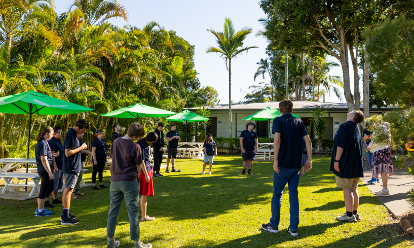 Participants and trainers stand in a large circle on a grassy outdoor area, taking part in a group stretching activity surrounded by palm trees and picnic seating.