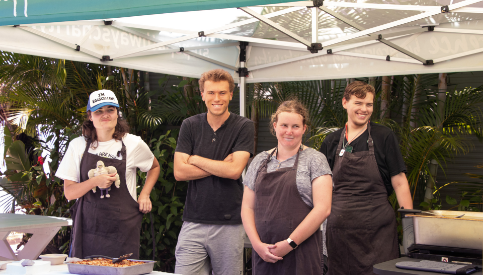 Four STEPS Pathways College participants stand behind a barbecue setup under a marquee, wearing aprons and helping prepare and serve food during a practical learning activity.