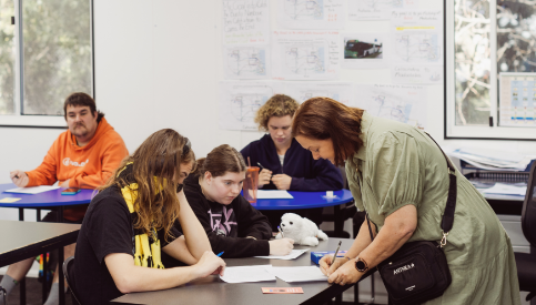 A trainer provides one-on-one support to participants working at desks in a classroom. Several participants are focused on written activities, while educational posters and notes are displayed on the classroom walls.