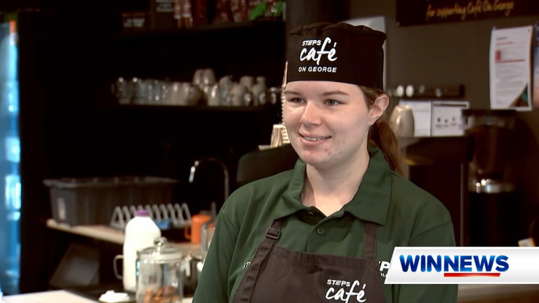 A young woman standing in a cafe, smiling at someone slightly off-camera. The 'WIN NEWS' logo is in the bottom right corner.