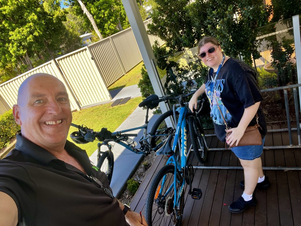 A man takes a selfie with a young woman. They are standing in front of a bike rack with two bikes parked on a shaded deck.