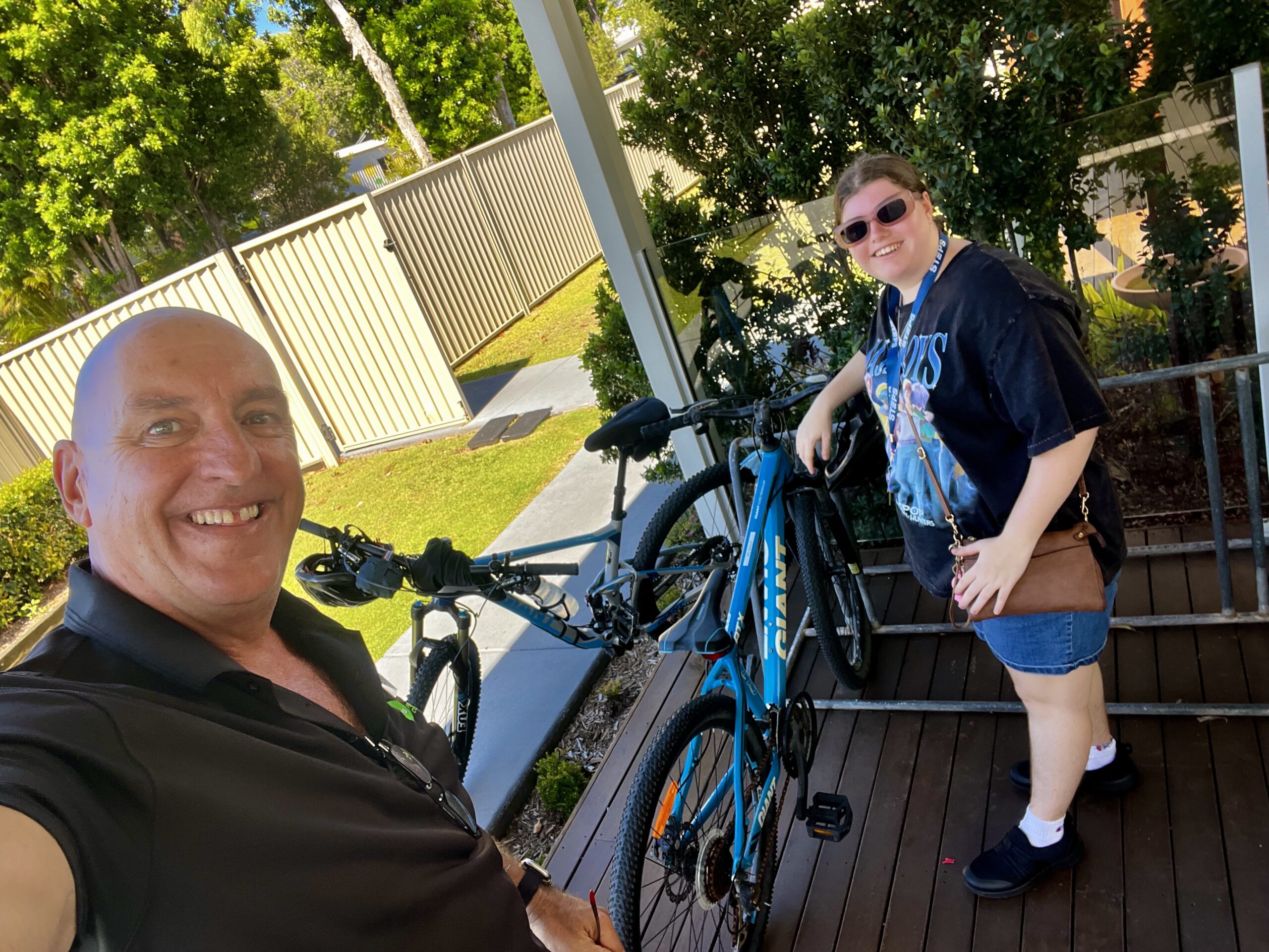 A man takes a selfie with a young woman. They are standing in front of a bike rack with two bikes parked on a shaded deck.