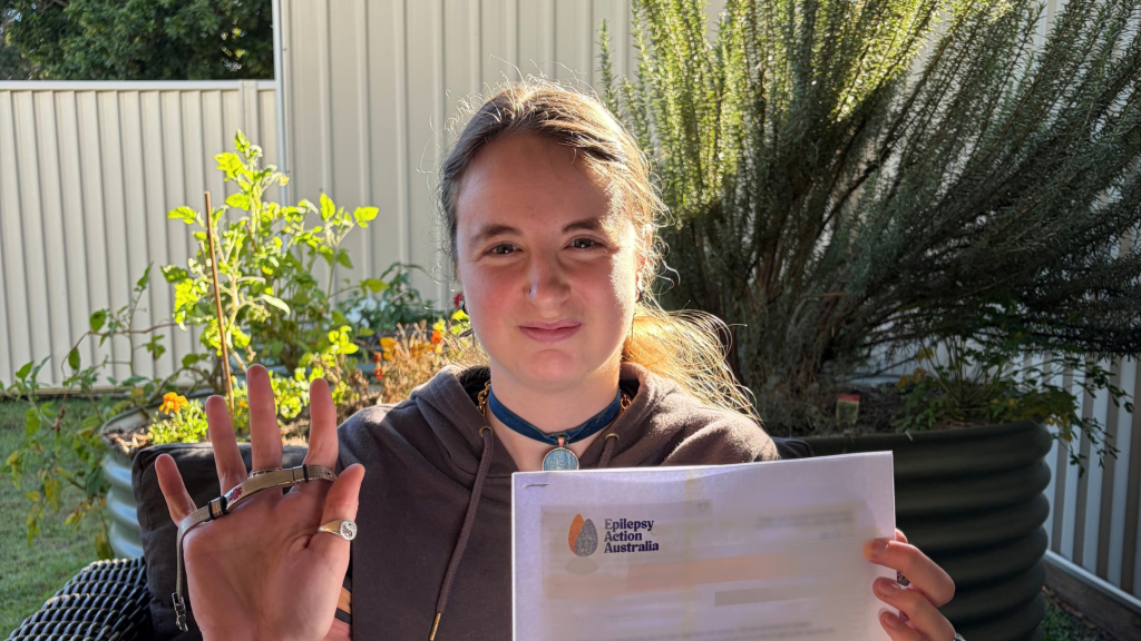 A young woman is smiling at the camera, holding a sheet of paper reading 'Epilepsy Action Australia' in one hand and a medical bracelet in the other. She is sitting outside on a sunny day at STEPS Pathways College.