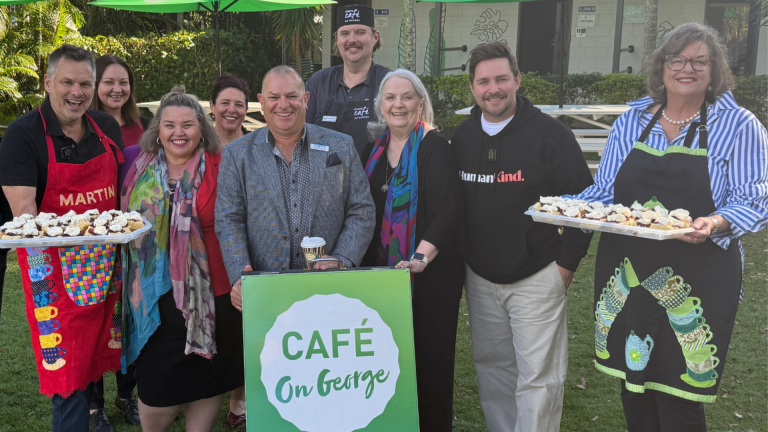 A group of people standing outside on a green lawn on a sunny day, with a sign in front of them reading ‘STEPS Cafe on George – can you smell the coffee yet?’ Two people are holding trays full of scones.