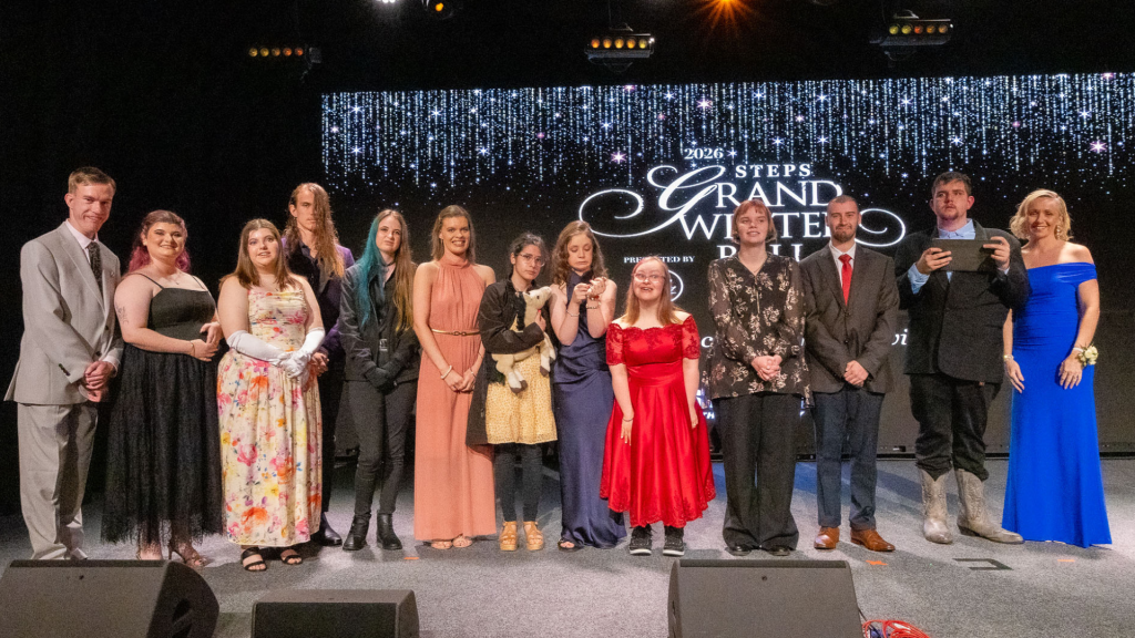 Thirteen people in formal wear stand on stage, smiling at the camera. Behind them is a screen that reads '2026 STEPS Grand Winter Ball.'