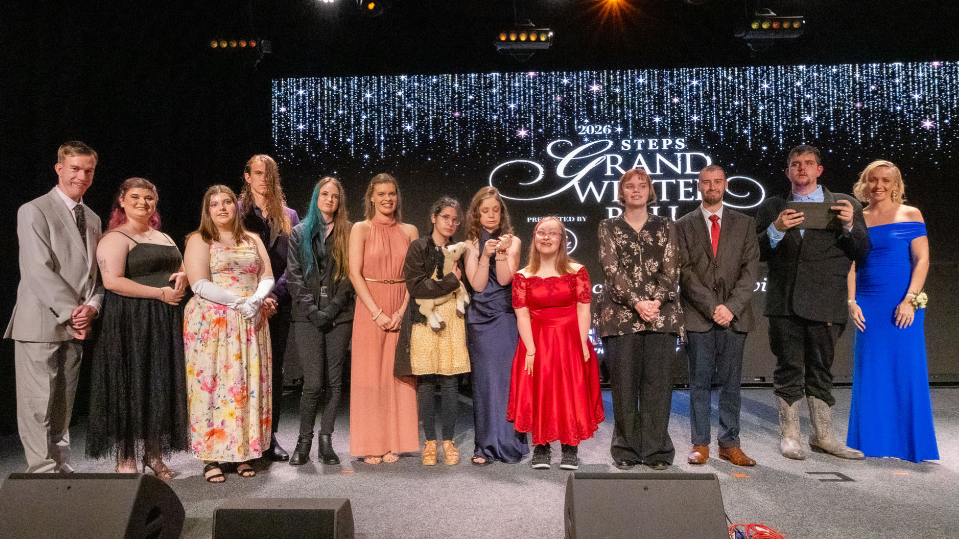 Thirteen people in formal wear stand on stage, smiling at the camera. Behind them is a screen that reads '2026 STEPS Grand Winter Ball.'