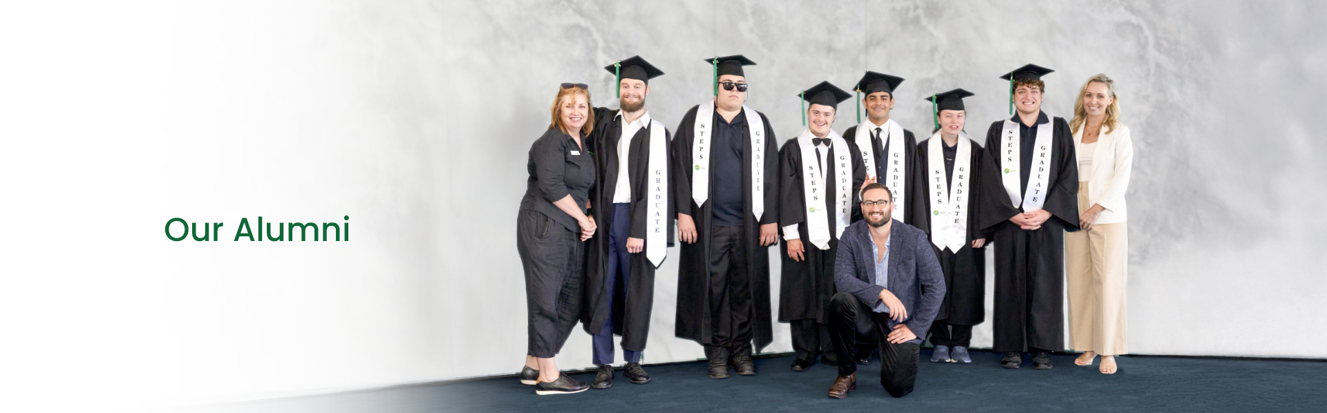 Group of STEPS Pathways College graduates wearing graduation gowns and mortarboards, posing with staff members at a graduation ceremony beneath the heading "Our Alumni".